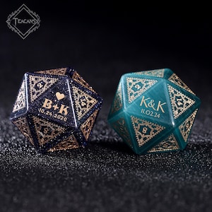 May include: Two personalized, faceted dice. One is dark blue with gold filigree and the initials "B+K" and the date "10.24.2020". The other is teal with gold filigree and the initials "K&K" and the date "11.02.24".