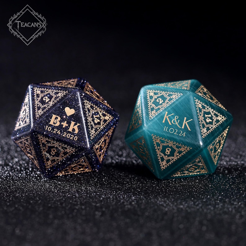 May include: Two personalized, faceted dice. One is dark blue with gold filigree and the initials "B+K" and the date "10.24.2020". The other is teal with gold filigree and the initials "K&K" and the date "11.02.24".