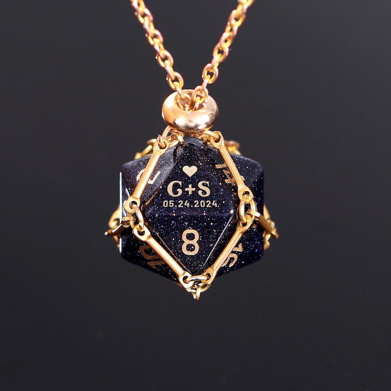 May include: A gold necklace with a blue, glittery, eight-sided dice pendant. The dice has gold numbers and a heart with the initials "G+S" and the date "05.24.2024".