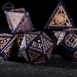 May include: A set of dark blue polyhedral dice with gold-colored intricate designs and glitter. One die features the initials "B/R" and the date "05.24.2024". The dice are for tabletop games.