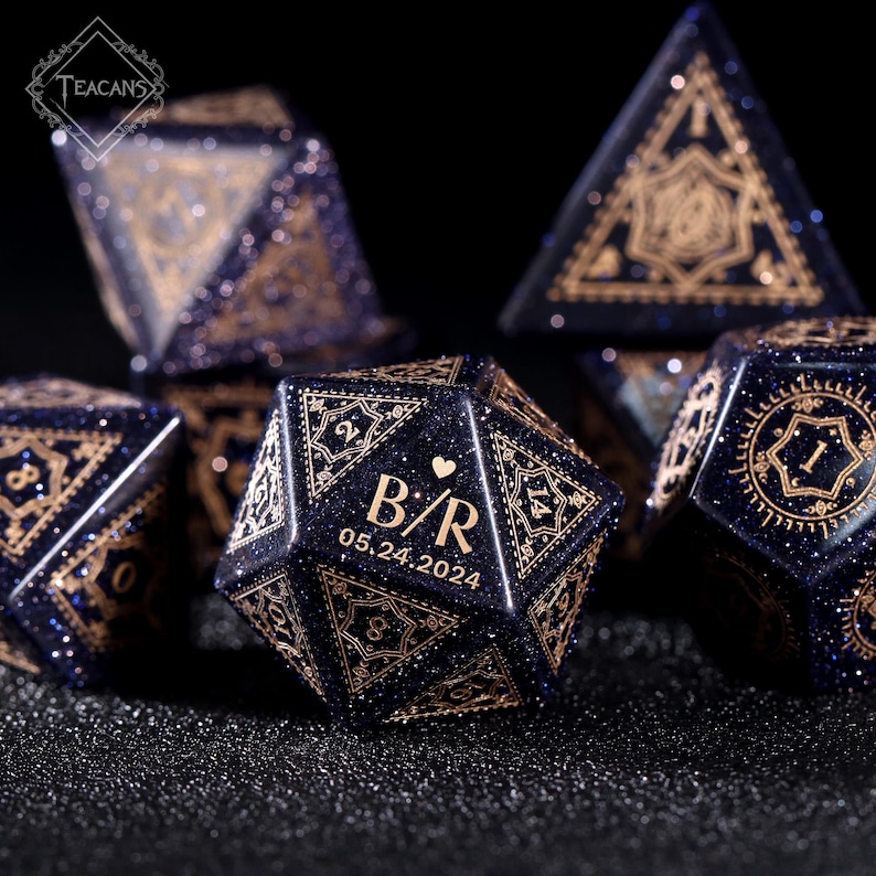 May include: A set of dark blue polyhedral dice with gold-colored intricate designs and glitter. One die features the initials "B/R" and the date "05.24.2024". The dice are for tabletop games.