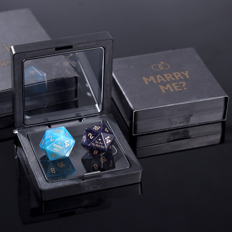 May include: Two blue and black gemstone dice with gold lettering in a black velvet box. The dice read "Marry Me?" and "You Are My Best Roll". The box has a black lid with the words "Marry Me?" in gold lettering.