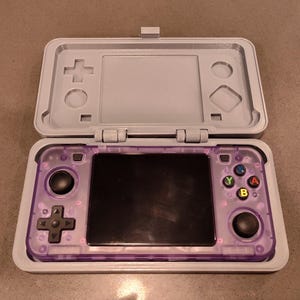 Puede incluir: Una consola de juegos portátil de color púrpura con una pantalla negra y botones. La consola está en una carcasa gris con una tapa con bisagras.