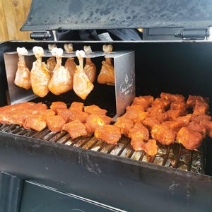 May include: A smoker grill with various cuts of seasoned meat. Chicken drumsticks hang from a rack, while ribs and cubed meat rest on the grill. The grill is black with a stainless steel rack.