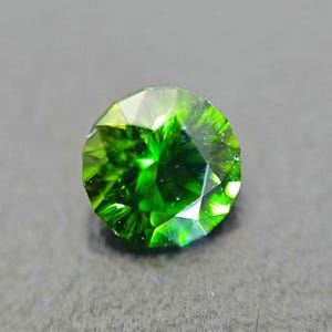May include: A round, faceted gemstone of a vivid, deep green hue. The gem is cut with multiple facets to reflect light, producing a sparkling effect. The gemstone is set against a grey background.