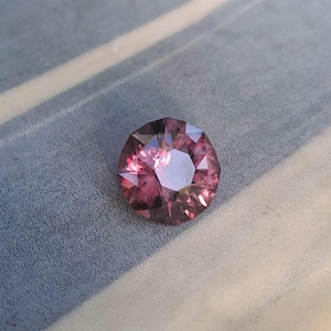 May include: A faceted, round gemstone with a deep pink hue. The gem's multiple facets reflect light, creating a sparkling effect. The gemstone is set against a textured, light blue surface.