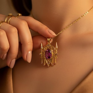 May include: A gold-colored pendant necklace featuring an ornate design with a large, rectangular, purple gemstone at the center. The pendant is accented with smaller pink gemstones and intricate filigree details. The necklace is displayed on a person.