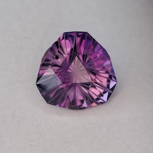 May include: A faceted, triangular gemstone with a deep purple hue. The gem's intricate cuts create a play of light, showcasing a gradient of colors from light to dark. The stone is set against a neutral background.