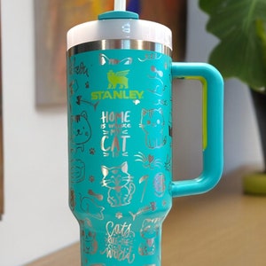 May include: A turquoise stainless steel travel mug with a black lid and a yellow handle. The mug has a silver design of cats and the text "Home is where the cat is". The mug is sitting on a wooden surface.