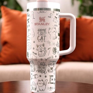 May include: White stainless steel travel mug with a pink lid and a black handle. The mug has a cat-themed design with the text "Home is where my cat is" and "Cats rule the world".