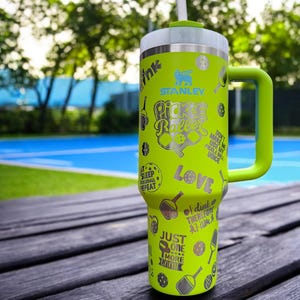 May include: A lime green stainless steel travel mug with a black handle. The mug has a variety of pickleball-themed designs and text, including "Pickleball", "Love", and "Just One More Game".