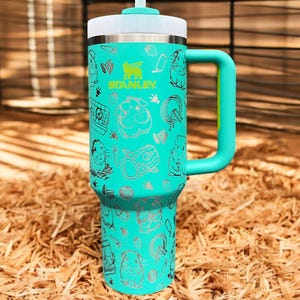 May include: A turquoise stainless steel travel mug with a black and white graphic print of a hamster and other small animal themed designs. The mug has a black lid with a straw and a black handle.