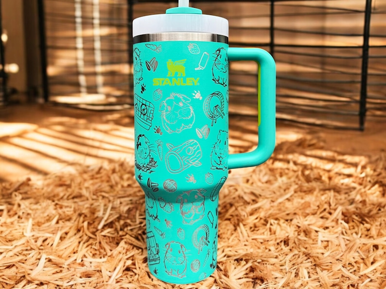 May include: A turquoise stainless steel travel mug with a black and white graphic print of a hamster and other small animal themed designs. The mug has a black lid with a straw and a black handle.
