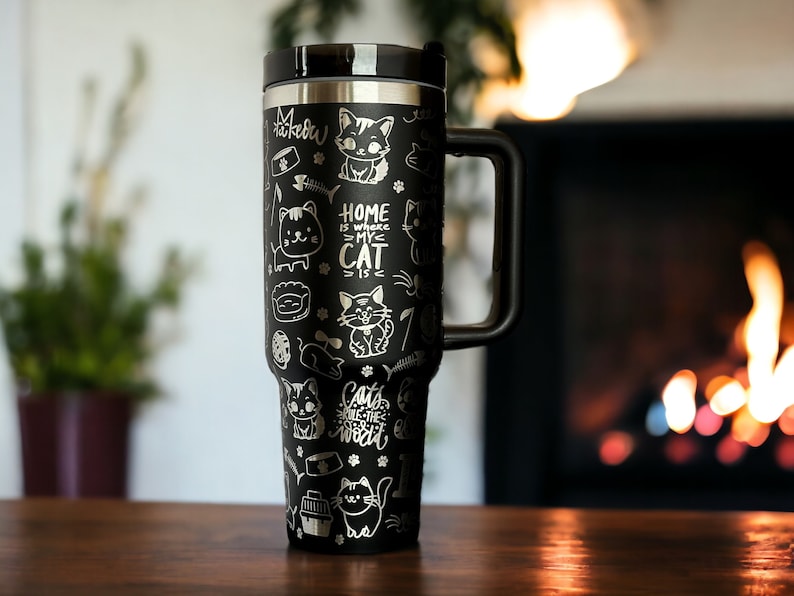 May include: Black stainless steel travel mug with a black handle. The mug is covered in a white design of cats, bones, and the text "Home is where my cat is".