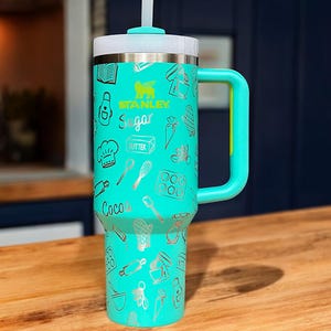 May include: A turquoise stainless steel travel mug with a black handle and a straw. The mug has a white lid and is decorated with a silver design of baking-themed illustrations, including the words "Stanley", "Sugar", "Butter", and "Cocoa".