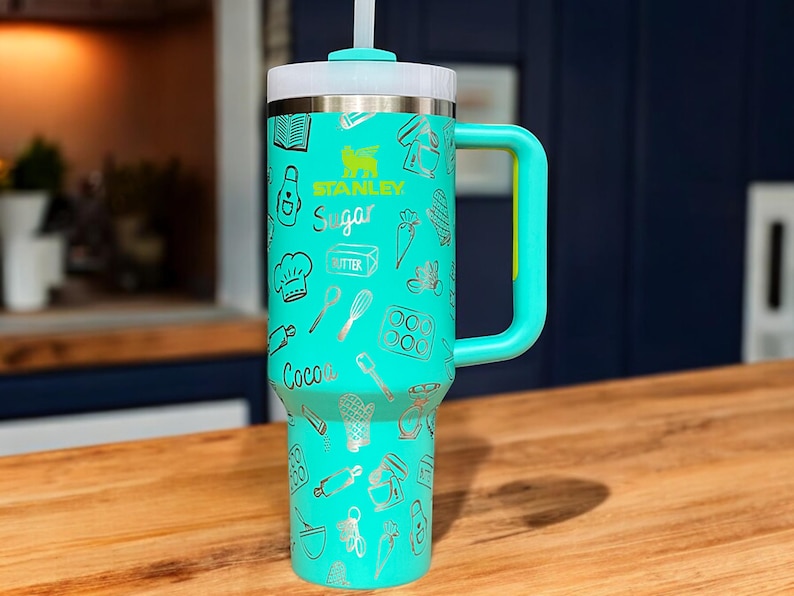May include: A turquoise stainless steel travel mug with a black handle and a straw. The mug has a white lid and is decorated with a silver design of baking-themed illustrations, including the words "Stanley", "Sugar", "Butter", and "Cocoa".