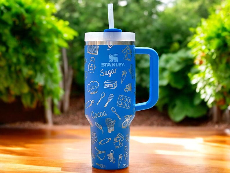 May include: A blue stainless steel travel mug with a handle and a straw. The mug has a white and silver design with the words "Stanley" and "Sugar" on it. The mug is decorated with a pattern of baking-related icons.