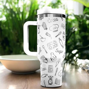 May include: White travel mug with a black and white pattern of baking-themed illustrations. The mug has a black lid and a black handle. The text "Cocoa" and "Sugar" are visible on the mug.