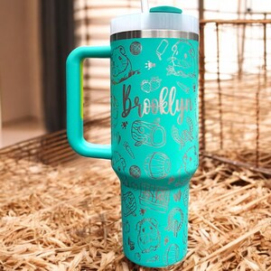 May include: Teal travel mug with a handle and a straw. The mug has a silver design of guinea pigs, carrots, and the name "Brooklyn".