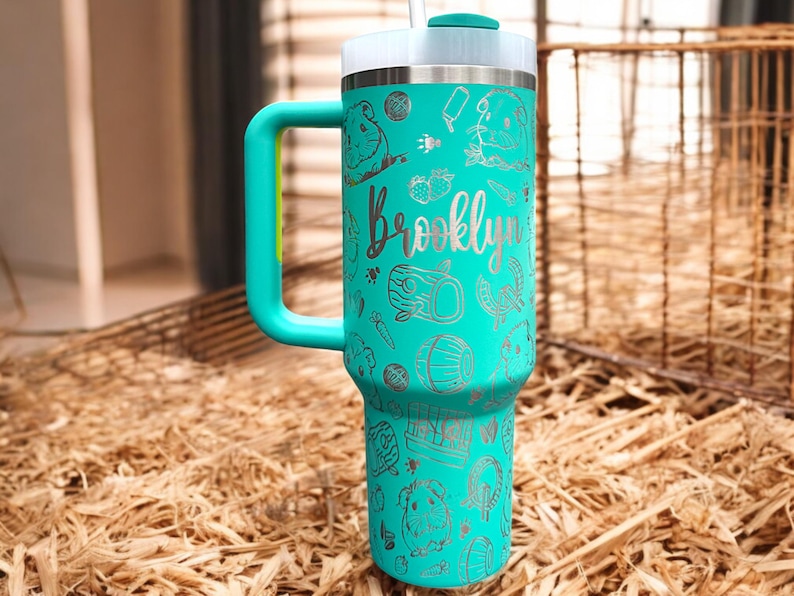May include: Teal travel mug with a handle and a straw. The mug has a silver design of guinea pigs, carrots, and the name "Brooklyn".