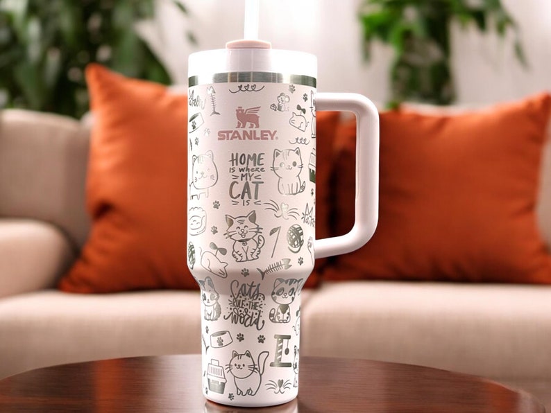 May include: White stainless steel travel mug with a pink lid and a black handle. The mug has a cat-themed design with the text "Home is where my cat is" and "Cats rule the world".