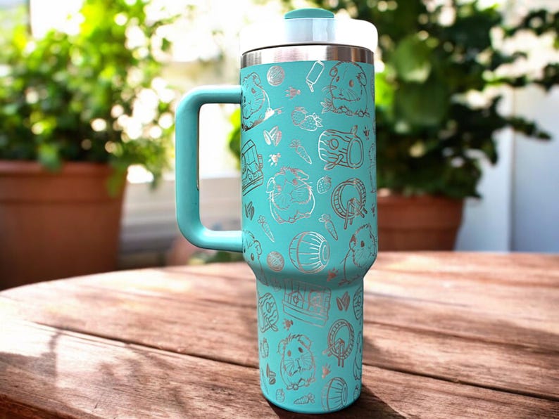 May include: A turquoise stainless steel travel mug with a handle. The mug is covered in a silver pattern of guinea pigs, hay, and other guinea pig-related items.