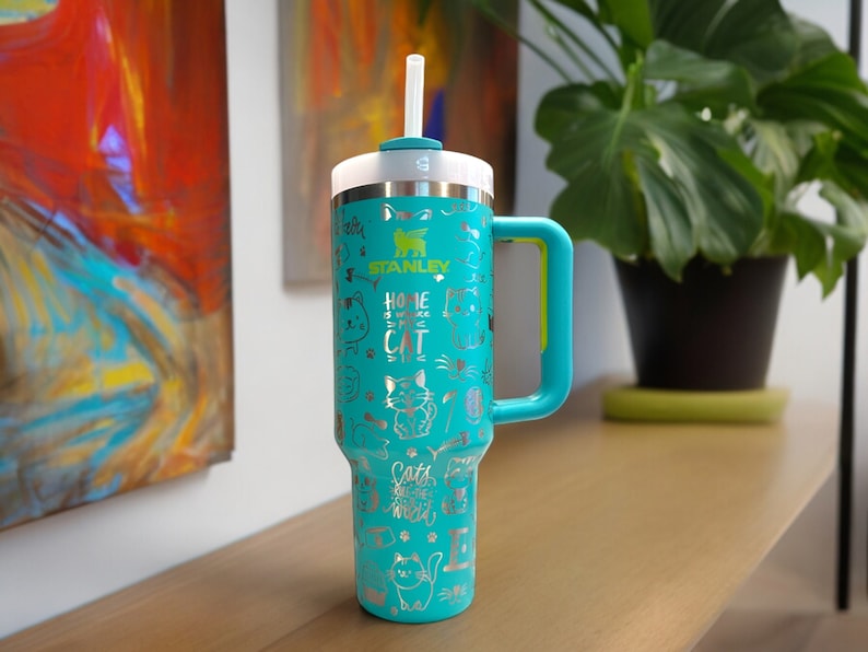 May include: A turquoise stainless steel travel mug with a black lid and a yellow handle. The mug has a silver design of cats and the text "Home is where the cat is". The mug is sitting on a wooden surface.