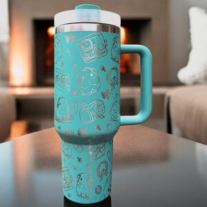 May include: A turquoise stainless steel travel mug with a silver lid and handle. The mug is decorated with a silver pattern of animals and trees.