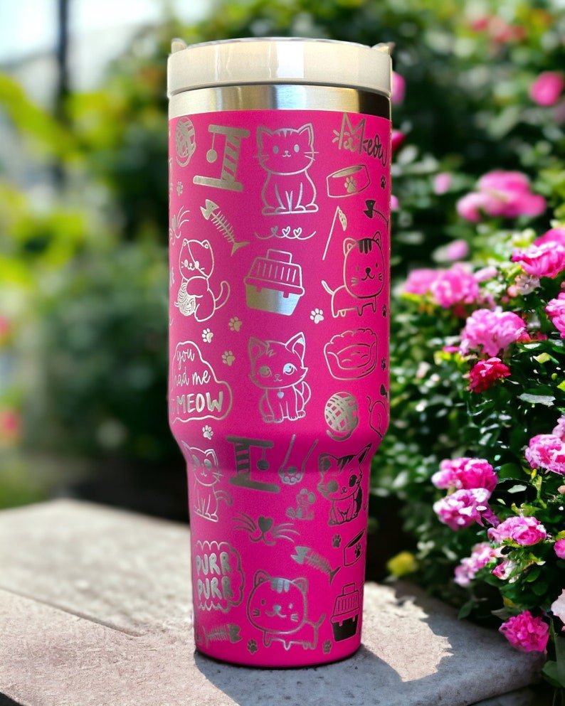 May include: A pink stainless steel tumbler with a silver cat-themed design. The tumbler features a variety of cat-related images, including cats, cat toys, and cat food bowls. The text "You had me at meow" is also included on the tumbler.