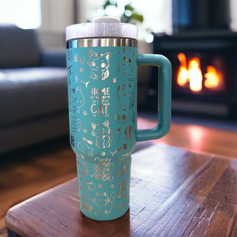 May include: A turquoise stainless steel travel mug with a silver lid and handle. The mug is covered in a silver cat-themed design with the text "Home is where my cat is" and "Cats rule the world".