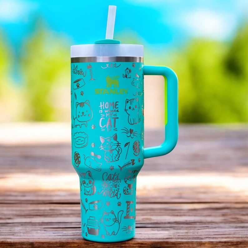 May include: A turquoise stainless steel travel mug with a black lid and a black handle. The mug has a cat-themed design with the words "Home is where my cat is" and "Cats rule the world".