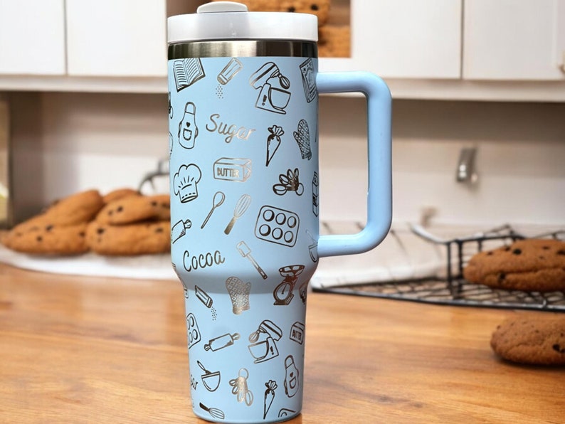 May include: A light blue stainless steel travel mug with a handle. The mug has a silver design of various baking-themed illustrations, including a whisk, rolling pin, and a chef's hat. The text "Cocoa" and "Sugar" are visible on the mug.