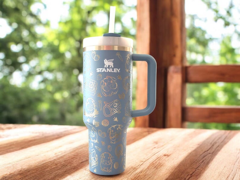 May include: A blue stainless steel travel mug with a handle and a straw. The mug has a gold design with a variety of images, including animals and plants. The mug is labeled "STANLEY".