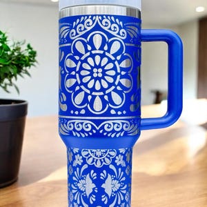 May include: A blue stainless steel travel mug with a silver floral design. The mug has a blue handle and a clear lid.