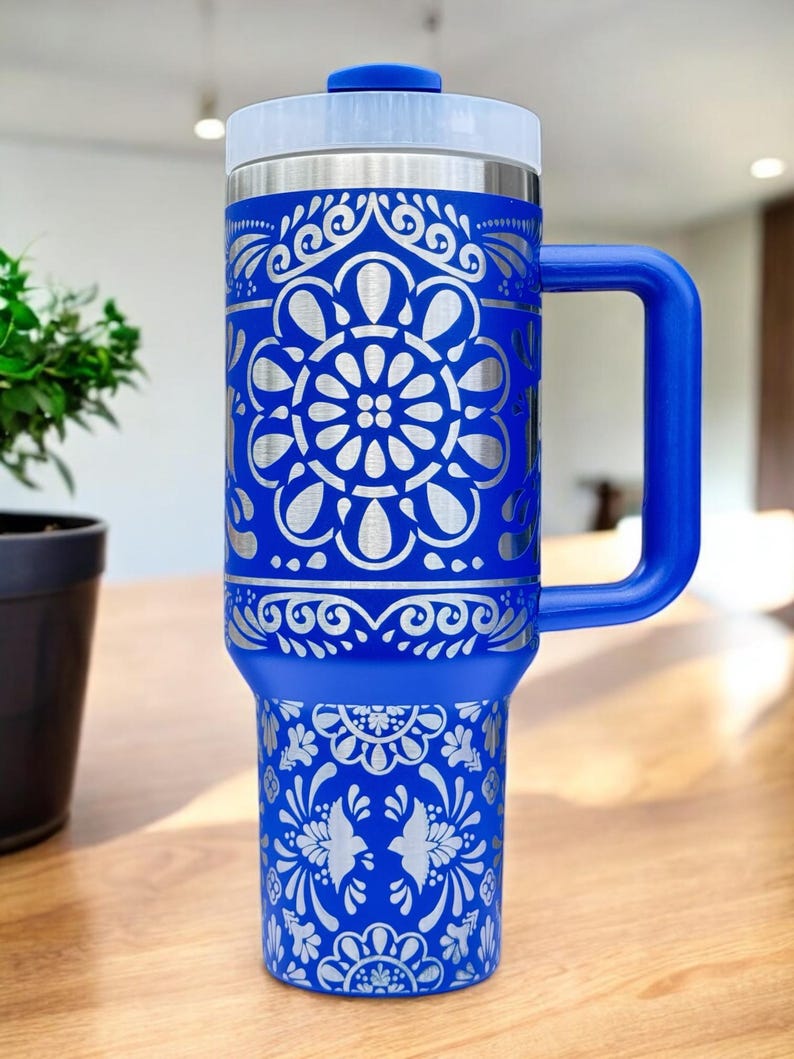 May include: A blue stainless steel travel mug with a silver floral design. The mug has a blue handle and a clear lid.