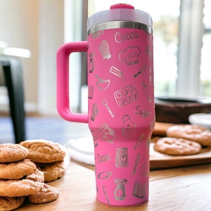 May include: Pink stainless steel travel mug with a handle. The mug has a silver design of baking tools and ingredients, including a whisk, rolling pin, and the words "Cocoa" and "Sugar".