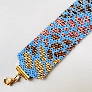 May include: A beaded bracelet with a blue background and a pattern of pink, brown, and gold beads. The bracelet has a gold-colored clasp. The bracelet is approximately 1.5 inches wide.