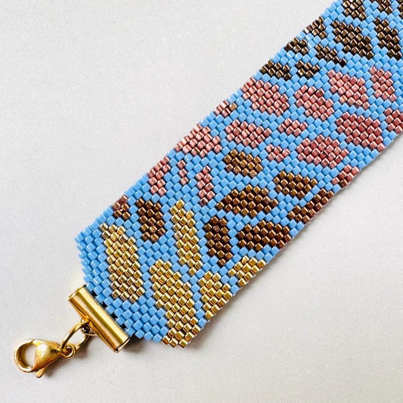 May include: A beaded bracelet with a blue background and a pattern of pink, brown, and gold beads. The bracelet has a gold-colored clasp. The bracelet is approximately 1.5 inches wide.