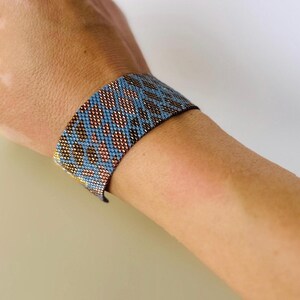 May include: A beaded bracelet in shades of blue, copper, and gold is worn on a wrist. The bracelet features a geometric pattern. The bracelet is approximately 1.5 inches wide.
