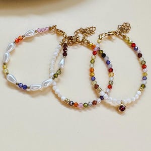 Puede incluir: Tres pulseras de oro con cuentas de colores y detalles de perlas blancas. Las pulseras presentan una variedad de colores, incluyendo rojo, naranja, amarillo, verde, azul, morado y rosa.