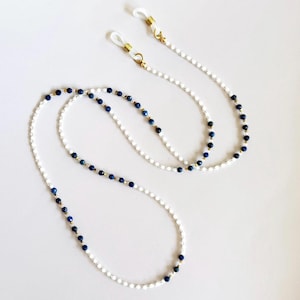 以下が含まれることがあります： 白とネイビーブルーのビーズとゴールドカラーのアクセントが特徴のメガネチェーン。メガネに取り付けるための透明なラバーループが付いています。メガネを安全かつアクセスしやすくするためのスタイリッシュなアクセサリーです。