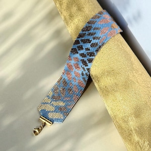 May include: A beaded bracelet with a blue, brown, and gold pattern. The bracelet has a gold-colored clasp. The bracelet is wrapped around a gold-colored cylindrical object.