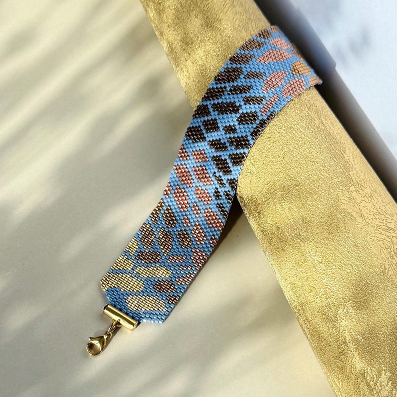 May include: A beaded bracelet with a blue, brown, and gold pattern. The bracelet has a gold-colored clasp. The bracelet is wrapped around a gold-colored cylindrical object.