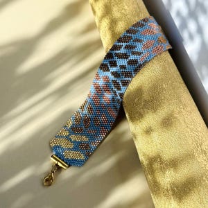 May include: A beaded bracelet with a blue, brown, and gold pattern. The bracelet has a gold-colored clasp. The design features a repeating pattern of small beads, creating a textured appearance. The bracelet is displayed on a gold-colored surface.