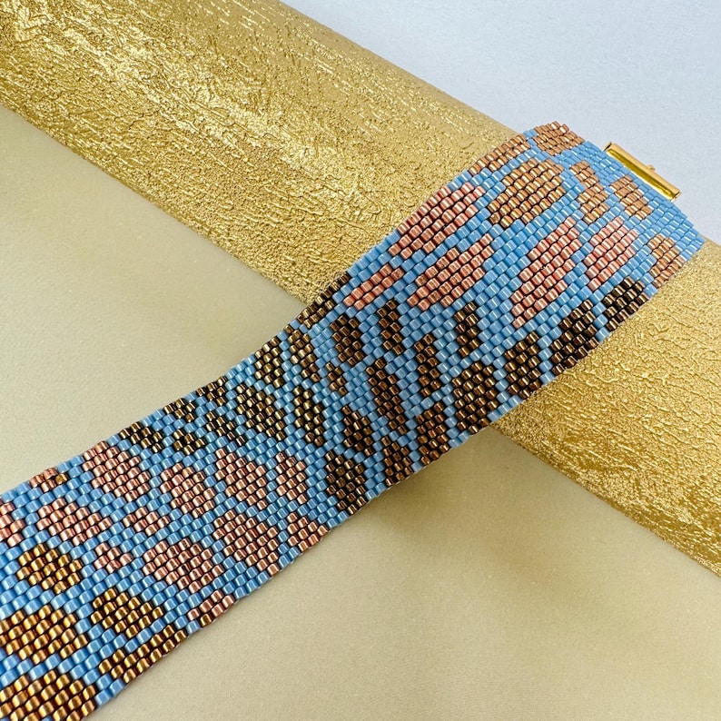 May include: A beaded bracelet with a geometric pattern. The bracelet features a light blue background with brown and pink diamond shapes. The clasp is gold-colored. The bracelet is approximately 1.5 inches wide.