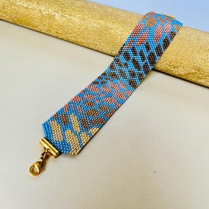 May include: A beaded bracelet with a blue, pink, and brown leopard print design. The bracelet has a gold-colored clasp and is displayed on a gold-colored surface. The bracelet is approximately 1.5 inches wide.