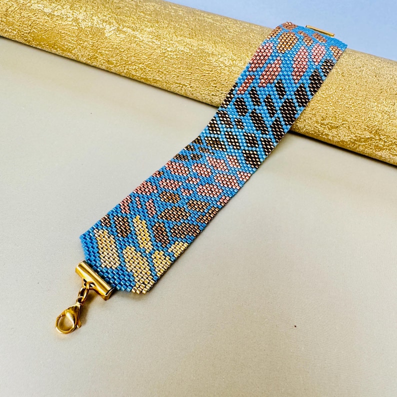 May include: A beaded bracelet with a blue, pink, and brown leopard print design. The bracelet has a gold-colored clasp and is displayed on a gold-colored surface. The bracelet is approximately 1.5 inches wide.