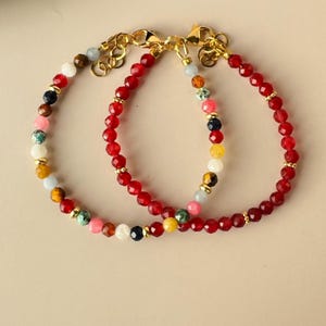 Op de afbeelding: Twee armbanden met kralen. Eén armband heeft rode facetkralen met gouden accenten. De andere armband heeft een mix van kleurrijke kralen, waaronder rood, geel, blauw en wit, met gouden accenten. Beide armbanden hebben gouden sluitingen.