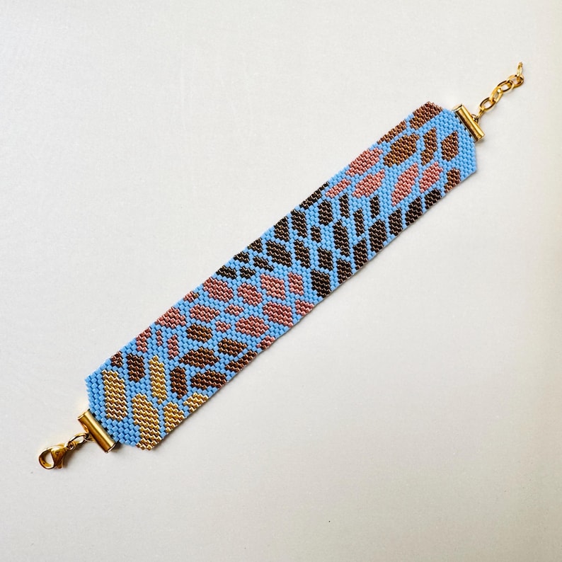 May include: A beaded bracelet with a blue background and a pattern of brown, pink, and gold shapes. The bracelet has gold-colored clasps and chain. The beaded design is a geometric pattern.