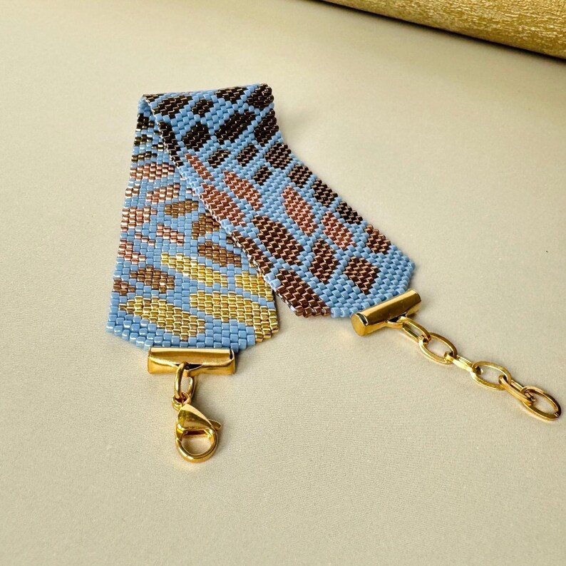 May include: A beaded bracelet with a geometric pattern in shades of blue, brown, gold, and copper. The bracelet has a gold-colored clasp and chain for closure. The woven design creates a textured appearance.